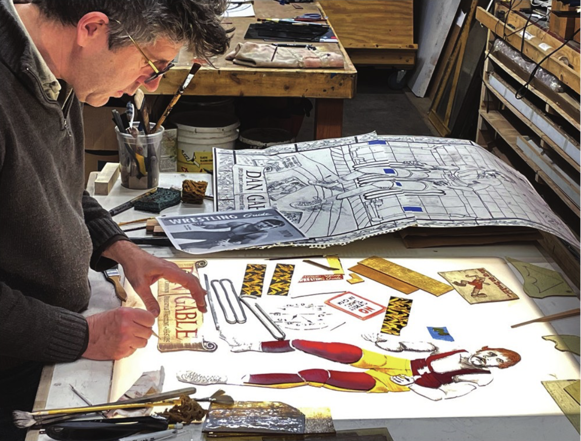 A person with gray hair and glasses looks down at a collage they are making. Their hands are positioned over the material moving it into position.